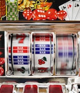 A close-up view of a slot machine with vibrant colors and various gambling symbols such as dice, coins, and playing cards. The reels display a combination of symbols, including bars, cherries, and the number seven. The chrome exterior and colorful details convey the theme of casino and gambling.