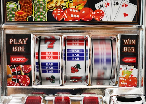 A close-up view of a slot machine with vibrant colors and various gambling symbols such as dice, coins, and playing cards. The reels display a combination of symbols, including bars, cherries, and the number seven. The chrome exterior and colorful details convey the theme of casino and gambling.