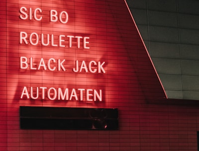 Red neon letters are mounted on a dark tiled wall, displaying the words Poker, Sic Bo, Roulette, Black Jack, and Automaten.