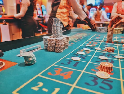 A casino scene with a roulette table features prominently, covered in green felt with colorful numbers and betting squares. Several stacks of poker chips are placed on different numbers, and the hands of several people are visible as they interact with the table. In the background, there are slot machines and more people, creating a vibrant and busy atmosphere.