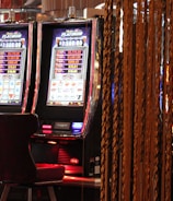 A pair of casino slot machines, each with detailed screens displaying various game outcomes and jackpot amounts. The chairs in front of the slot machines are dark and cushioned for comfort. The background features a hanging curtain made of golden ropes and other casino decorations are visible.