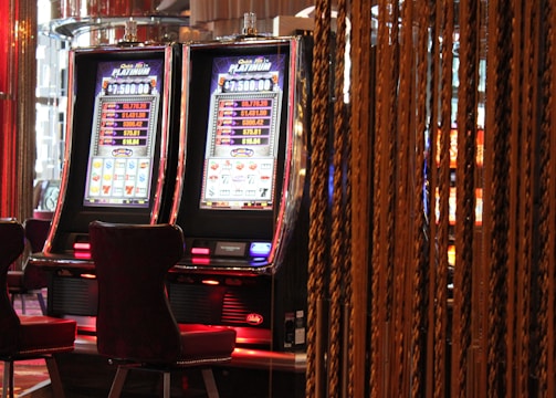 A pair of casino slot machines, each with detailed screens displaying various game outcomes and jackpot amounts. The chairs in front of the slot machines are dark and cushioned for comfort. The background features a hanging curtain made of golden ropes and other casino decorations are visible.