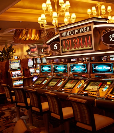 A casino floor featuring rows of brightly lit video poker machines. The area is illuminated by elegant chandeliers, with a display board showing a large sum of money, suggesting a possible jackpot. The rich decor includes luxurious carpets and comfortable seating, adding to the opulent atmosphere.