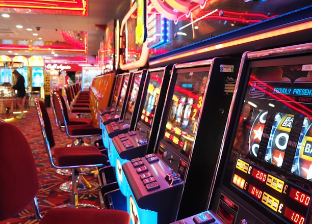 A brightly lit casino interior featuring a row of colorful slot machines. The neon lights create a vibrant atmosphere, with patterned carpeting adding to the decor. Stools are aligned neatly in front of each machine.