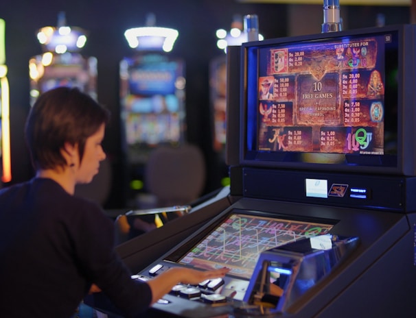 A person is seated at a slot machine console in a casino, focusing intently on the game displayed on the screen. The background shows several other illuminated gaming machines, adding a vibrant and colorful atmosphere to the scene.