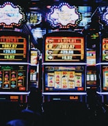 A row of colorful slot machines lit with bright neon lights, showcasing various graphical elements on the screens, indicating different games and jackpots. The atmosphere suggests a lively casino setting, bustling with excitement.