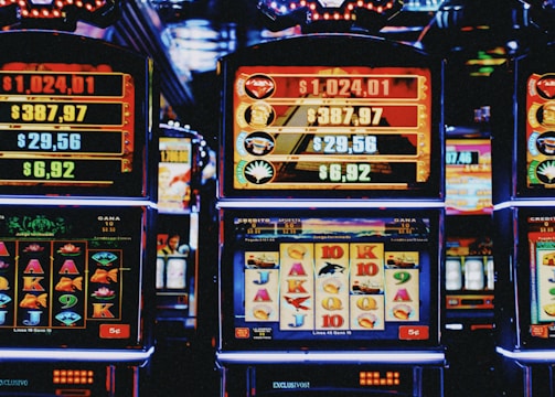 A row of colorful slot machines lit with bright neon lights, showcasing various graphical elements on the screens, indicating different games and jackpots. The atmosphere suggests a lively casino setting, bustling with excitement.