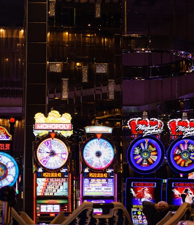 A lively casino scene featuring a row of bright, colorful slot machines with themes like Monopoly, Michael Jackson, and Lord of the Rings. The gaming machines display a variety of flashing lights and digital screens, creating an energetic and exciting atmosphere. Reflections of the lights are visible in glossy surfaces above the machines.