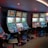 A row of brightly lit slot machines in a casino setting, each with colorful displays and various game themes. The machines are lined up against a dark wall and are unoccupied, with cushioned chairs placed in front of them. The area is dimly lit except for the glow from the screens, creating a vibrant and engaging atmosphere.