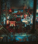 A bustling casino floor filled with groups of slot machines and people walking around. The atmosphere is vibrant with neon lights and illuminated signs. The interior has a ceiling supported by large pillars and there are stairs leading down to the gaming area.