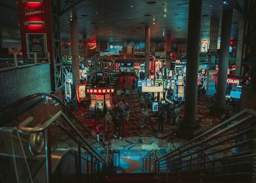 A bustling casino floor filled with groups of slot machines and people walking around. The atmosphere is vibrant with neon lights and illuminated signs. The interior has a ceiling supported by large pillars and there are stairs leading down to the gaming area.