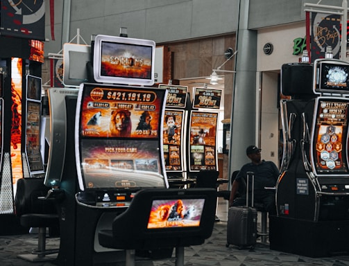 A collection of slot machines is situated inside a building with bright, colorful screens displaying various games and gaming themes. A person is seated nearby, seemingly relaxed with luggage beside them, and the atmosphere is lively with electronic displays.